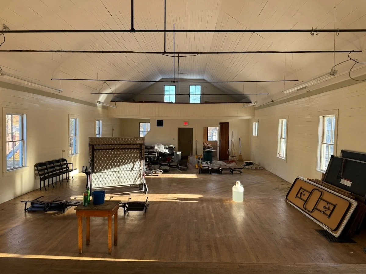 Empty large event hall or community center with wooden floor, several windows, chairs, tables, and folding tables pushed to the side, with some supplies and equipment scattered around.