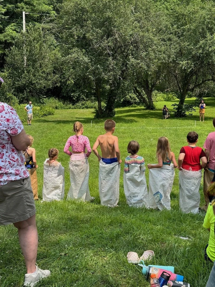 Children and adults participating in a sack race outdoors on a grassy field with trees, under a sunny sky.