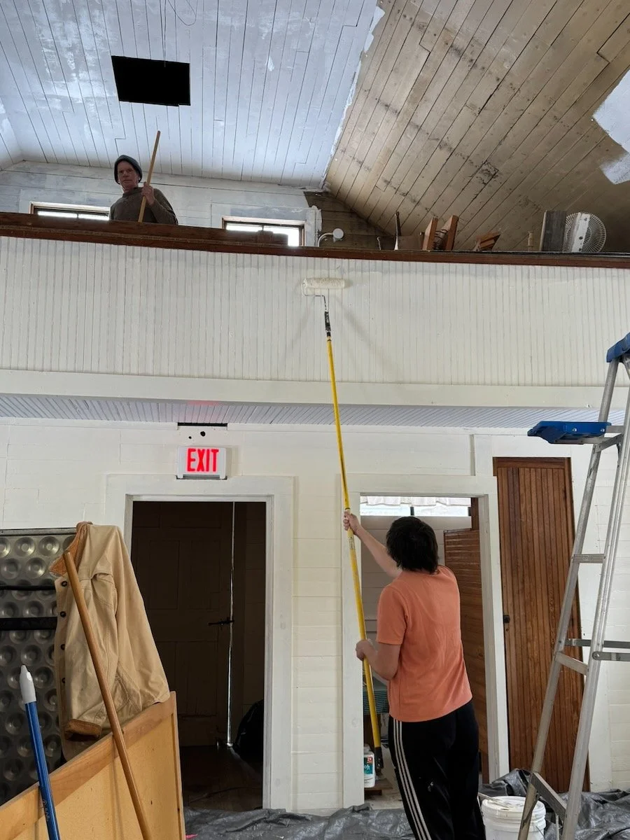 Two people painting an interior room, one using a roller on an extension pole and the other standing on a ladder. The room has a white wall, a wooden ceiling, and an upper level with windows and a person holding a long stick. Blue painter's tape, a l