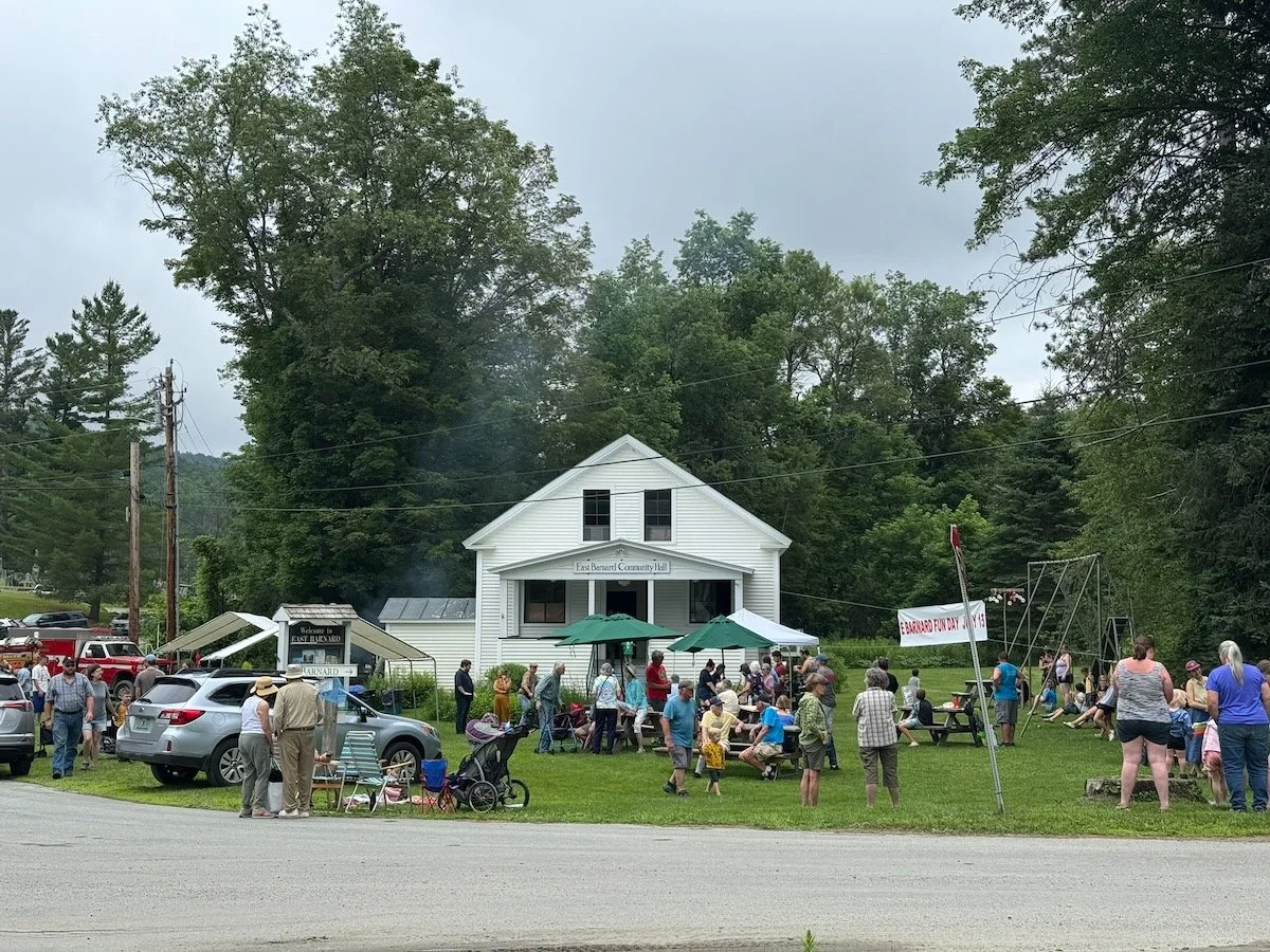 A community gathering in front of a white building with a sign that reads 'East Barnard Community Hall.' People are socializing, some sitting under umbrellas, and there are children playing on swings. There are several cars parked nearby and a banner