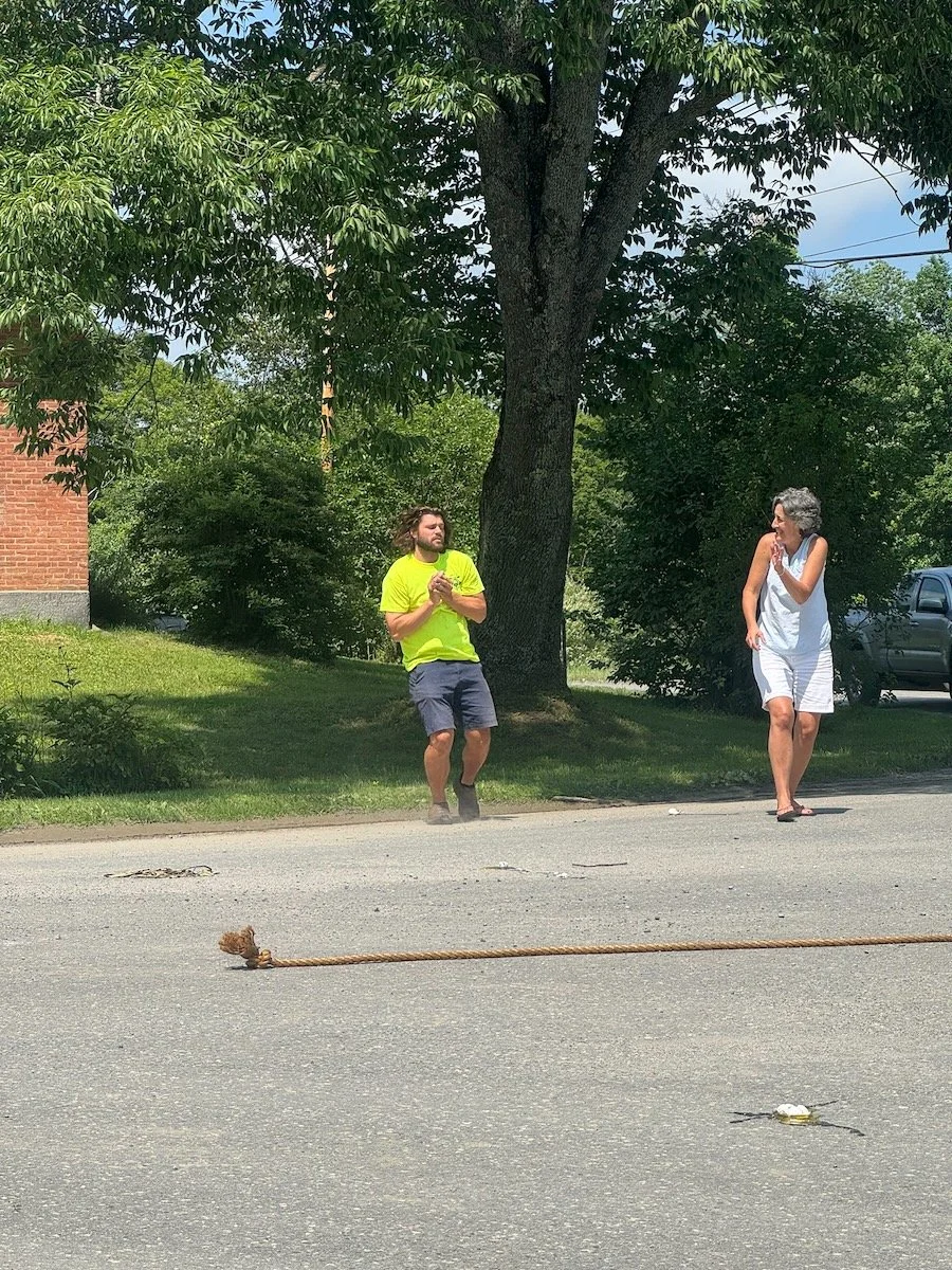 A man and a woman standing on the sidewalk next to a large tree, with a rope tied to the ground in front of them. The man is wearing a bright yellow shirt and dark shorts, and the woman is dressed in white. It is a sunny day in a green, leafy area.