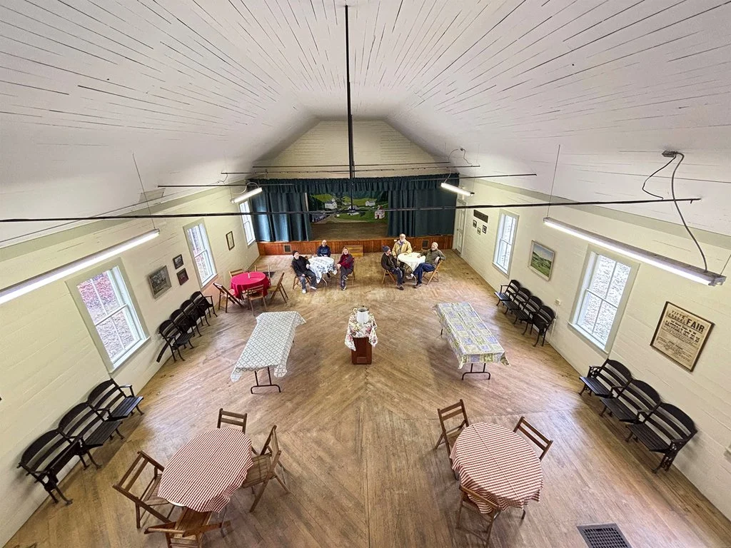 A community hall with a stage, tables, chairs, and people sitting and conversing.