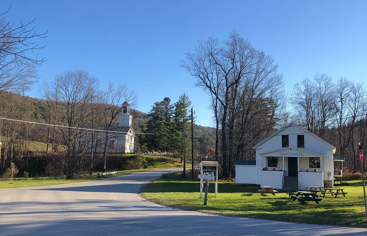 A small white community hall with a sign reading 'East Burnard Community Hall' and multiple outdoor picnic tables in front. A church with a steeple is visible on a hill in the background, along with leafless trees, a clear blue sky, and a winding road leading up to the buildings.