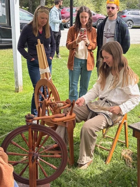 A woman sitting on a wooden chair demonstrating traditional spinning with a spinning wheel, while four people watch nearby at an outdoor event.