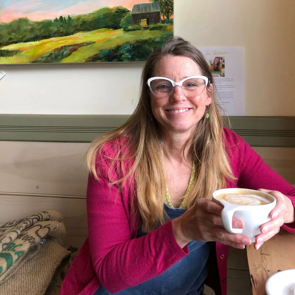 Sue Schlabach, East Barnard Crier. A woman with long hair, glasses, and a bright smile wearing a pink cardigan and green earrings, holding a cup of coffee, sitting in a cozy cafe with a landscape painting on the wall behind her.