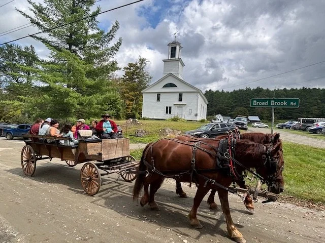 A horse-drawn wagon with several people seated, passing by a white church with a steeple, parked cars, and a green road sign that reads 'Broad Brook Rd' on a cloudy day.