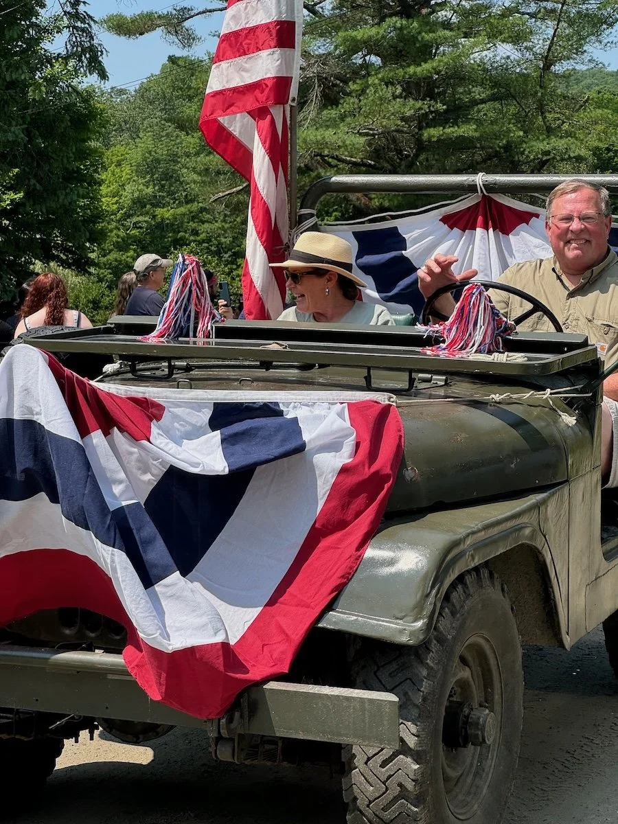People riding in a military vehicle decorated with American flags and bunting, celebrating a patriotic event outdoors.