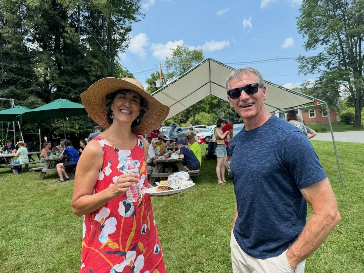 A woman wearing a large sun hat and a colorful dress, holding a plate of food and a bottle of water, stands next to a man in sunglasses and a dark blue T-shirt, with tables and people in the background at an outdoor gathering under a tent on a grassy