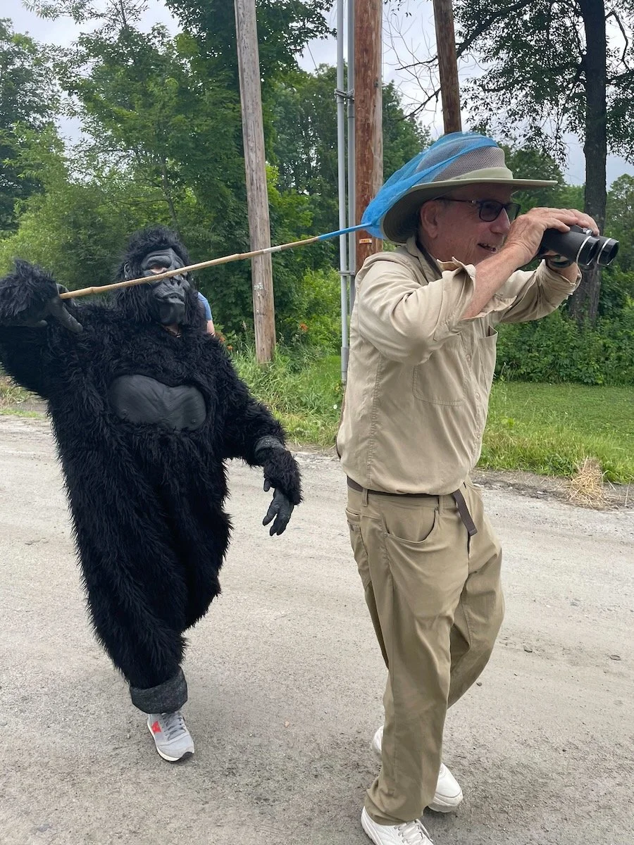 A man dressed as a safari explorer is walking while holding binoculars and wearing a net hat. A person in a gorilla costume is walking behind him, holding a stick that is resting on the explorer's shoulder. The scene is outdoors with green trees and 