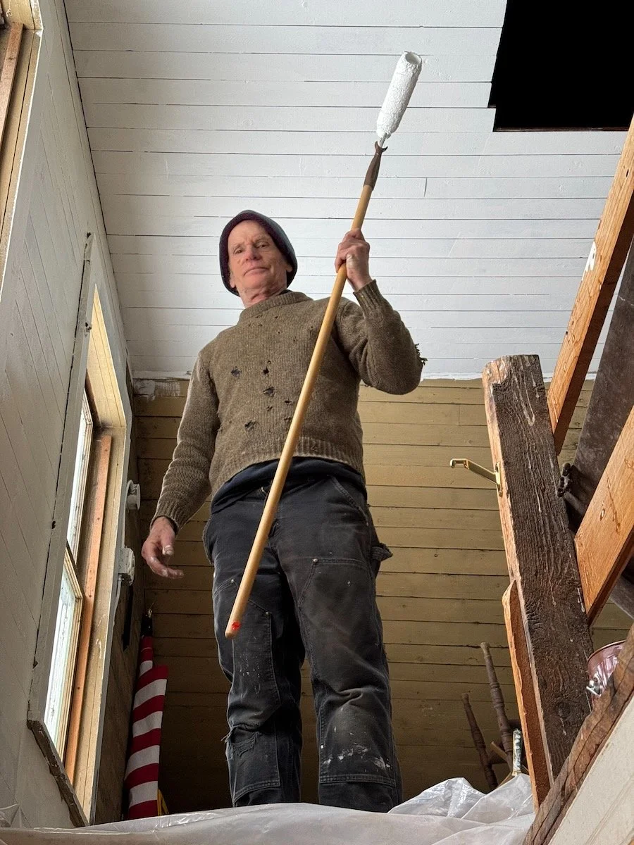 A person standing on a ladder holding a paint roller, painting a ceiling in a room with wooden walls.