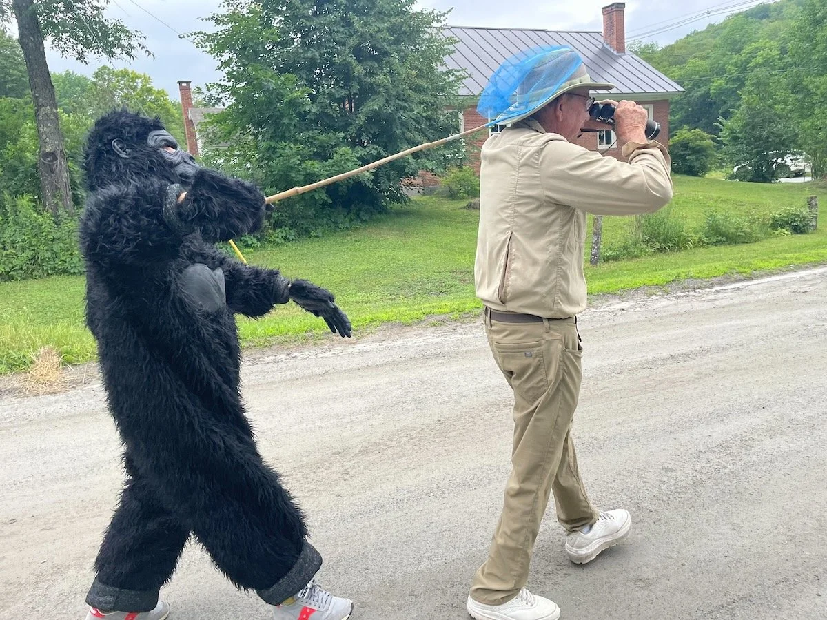 A person dressed in khaki clothing and a hat with a blue netting plays binoculars while a person in a gorilla costume, holding a stick like a leash, walks behind them on a gravel road with houses and green trees in the background.
