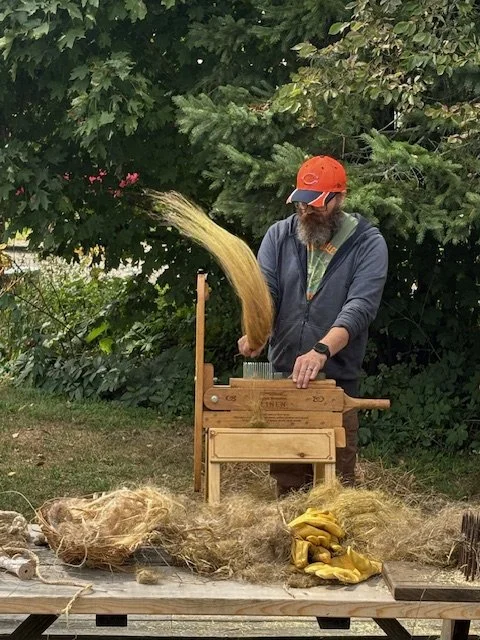 A man with a beard wearing a gray hoodie and orange cap using a flax combing tool, causing straw to fly into the air. The scene is outdoors with green trees and bushes in the background.