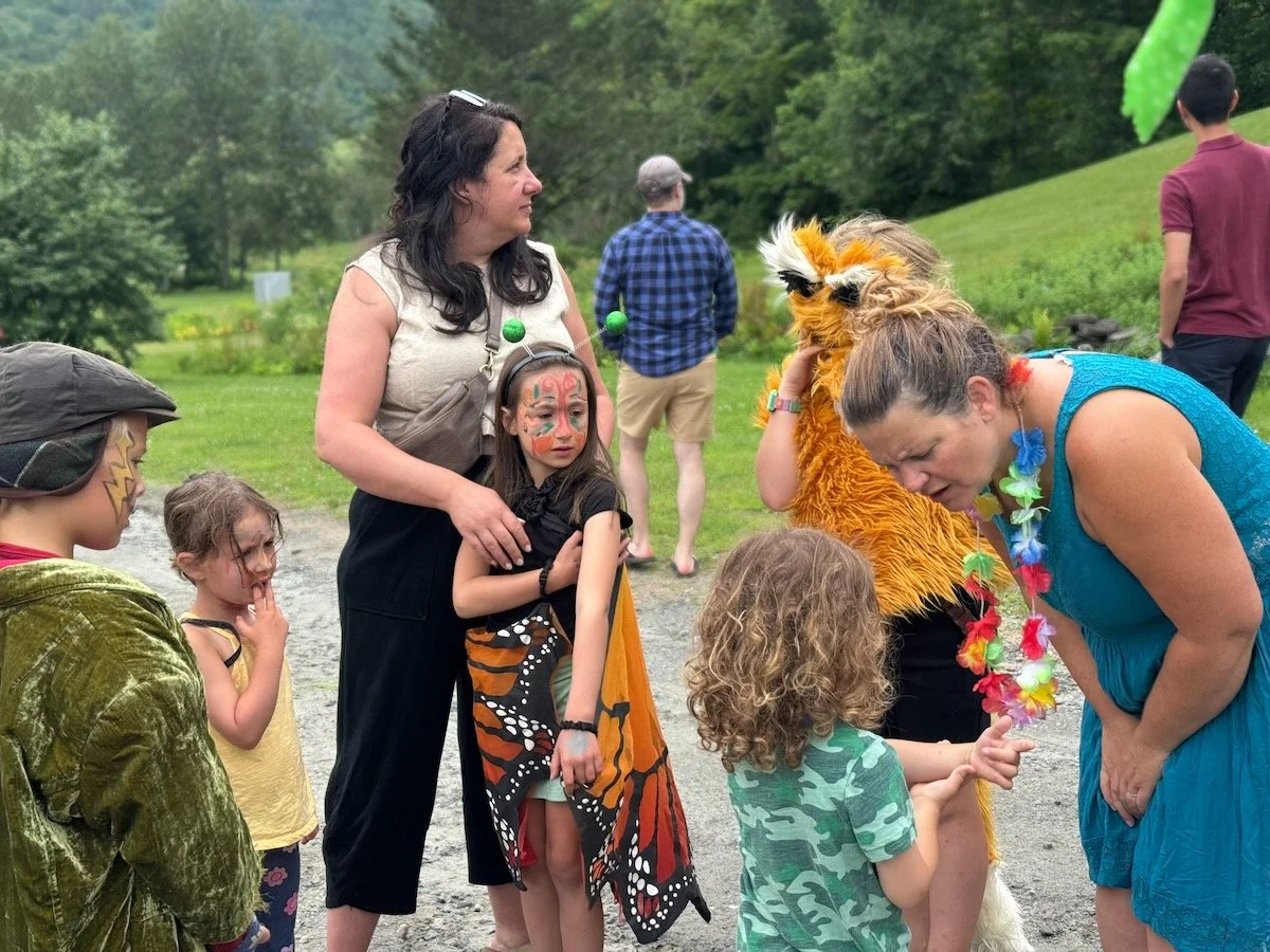 Children and adults dressed in colorful costumes, with face paint and accessories, gathered outdoors on a grassy area with trees in the background, during a festive event.