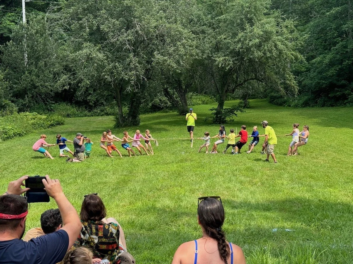 Children and adults participating in a tug-of-war game on a grassy field surrounded by trees, with spectators watching and capturing the event on their phones.