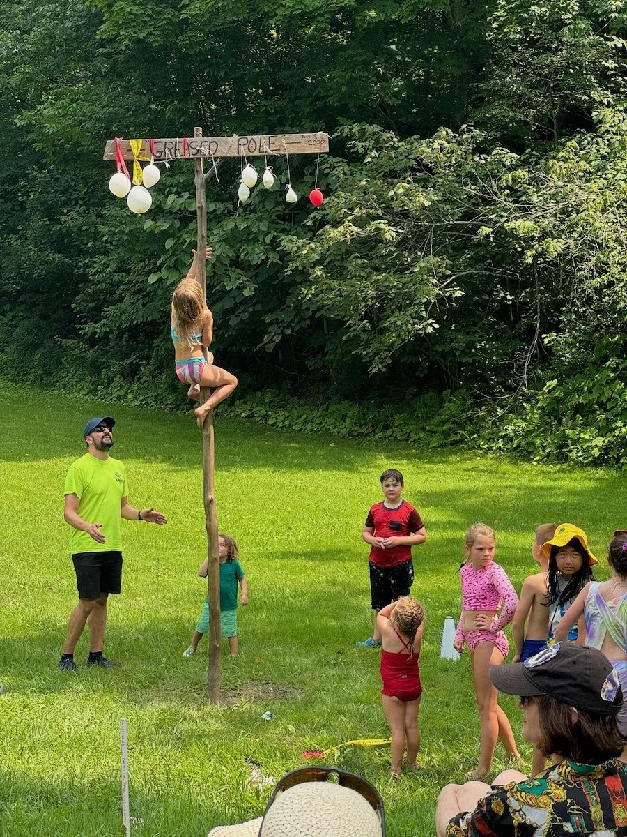 Children participate in a game of 'Grapevine Pole' at a summer outdoor gathering. A girl in a swimsuit is climbing a pole with balloons hanging from a wooden structure labeled 'Grapevine Pole.' Several other kids and adults stand around watching, som