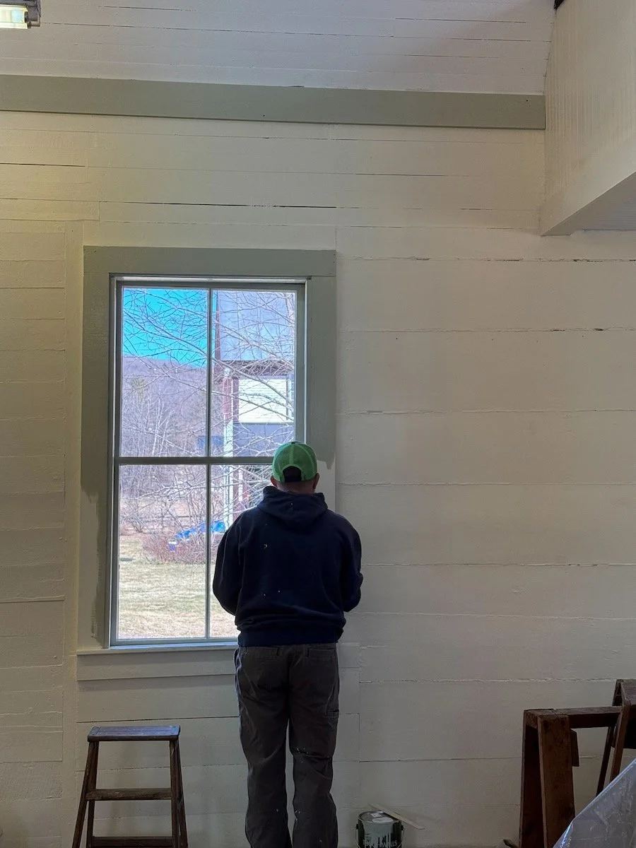 Man with a green baseball cap standing and looking out a window in a room under construction or renovation.