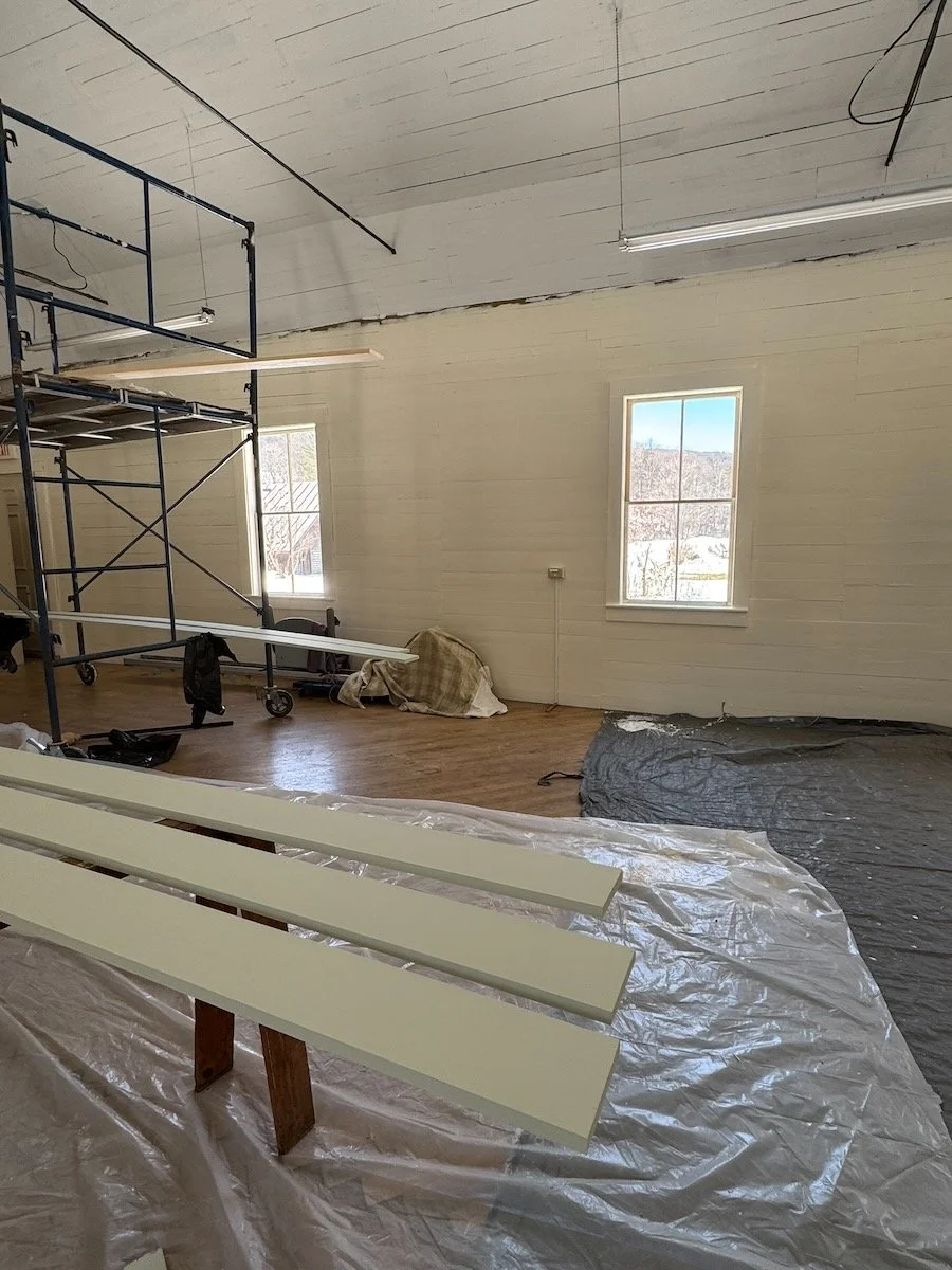 Interior of a room under renovation with white painted wooden walls, two windows, a ceiling with visible wiring, a metal scaffold, and a plastic sheet covering the floor, with some wooden slats placed on a makeshift stand.