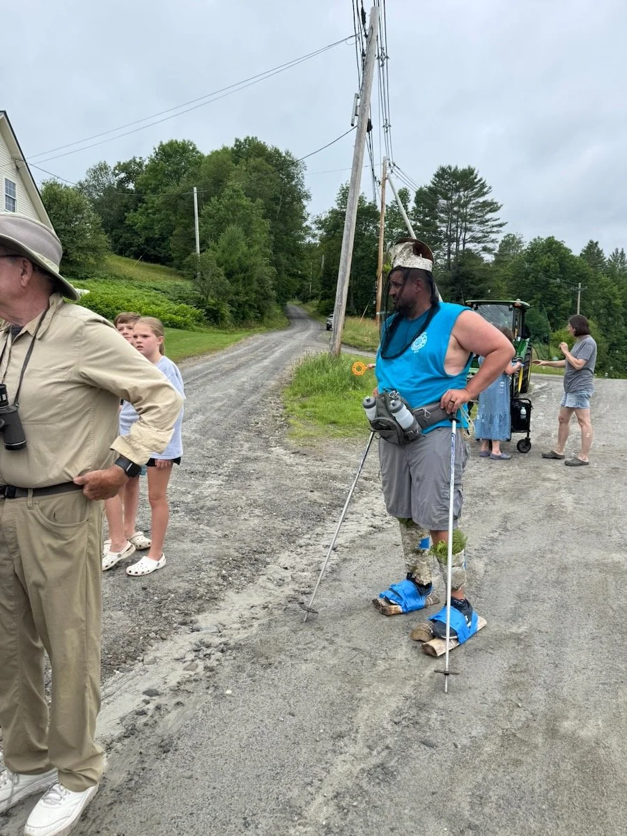 Man with prosthetic legs walking on a gravel road using makeshift wooden crutches, wearing a blue sleeveless shirt and shorts, surrounded by people, with trees and power lines in the background.