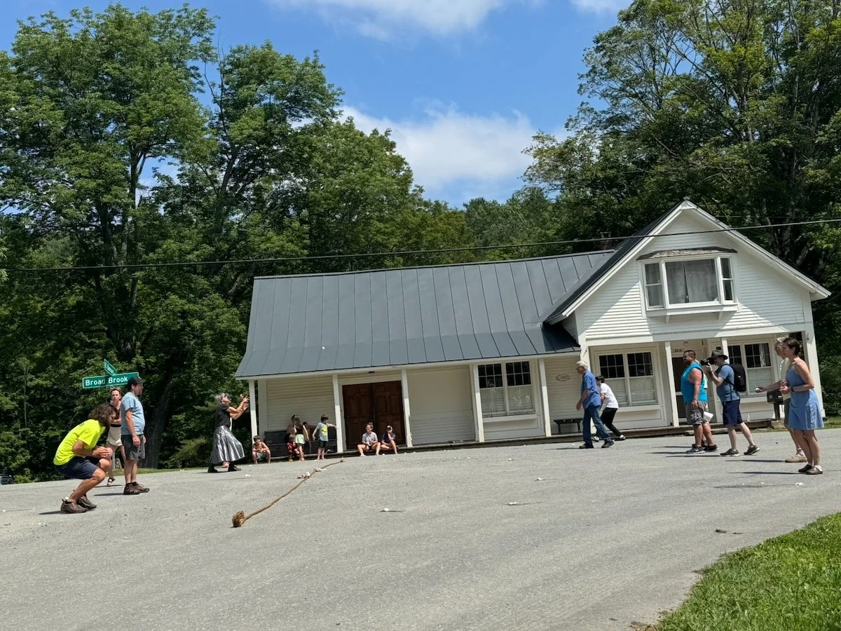 People playing a game outside a white house with a gray roof, on a sunny day with green trees in the background.
