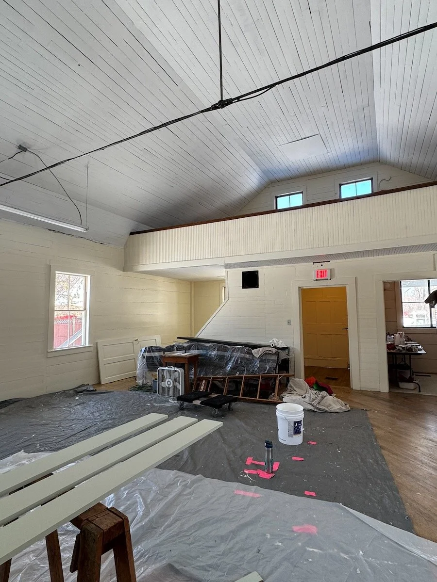 Interior of a house under renovation with plastic covering the floor, a white bench, construction tools, a fan, and supplies scattered around.
