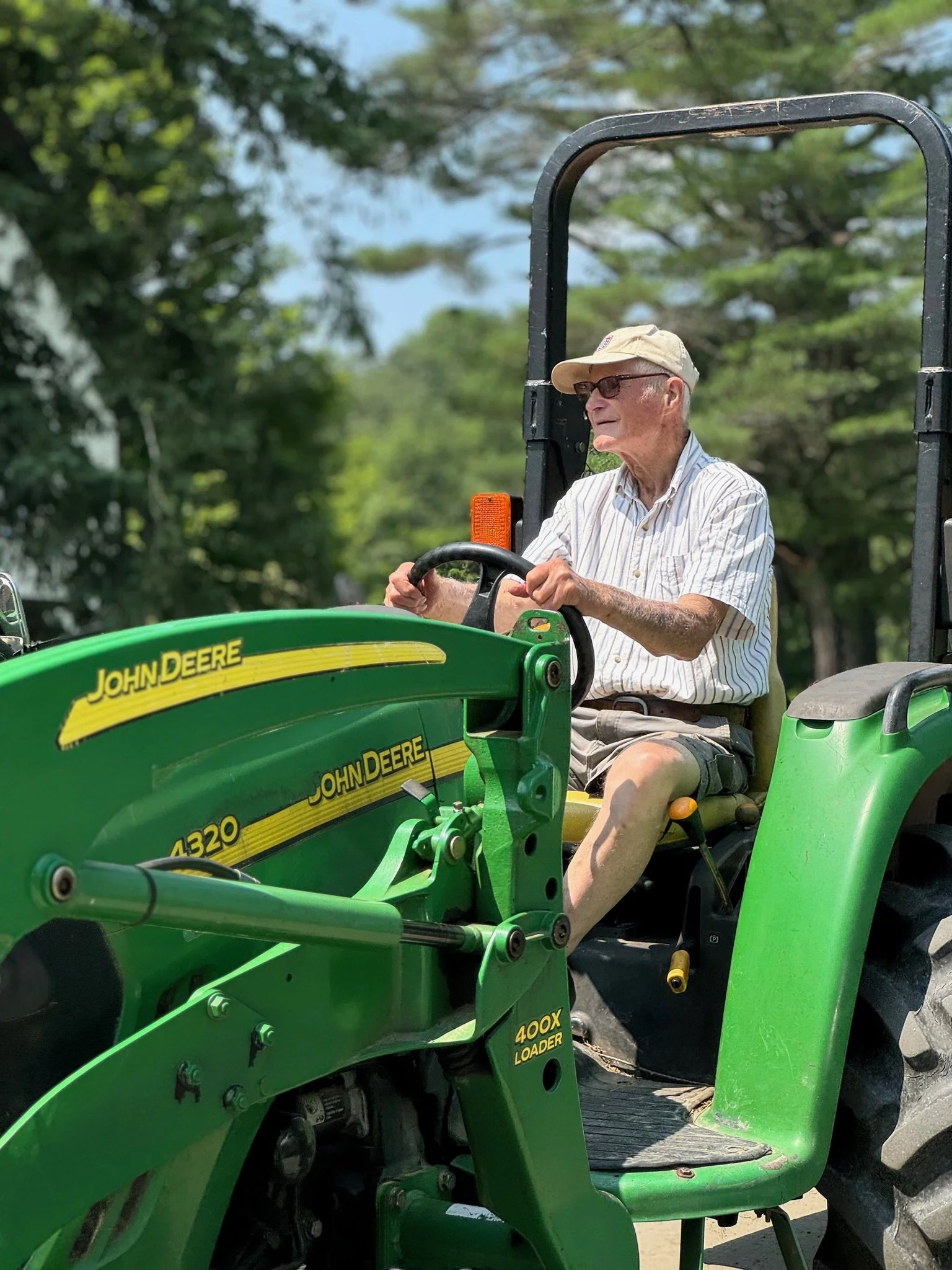 An elderly man wearing a beige cap glasses a striped shirt and shorts driving a green John Deere tractor.