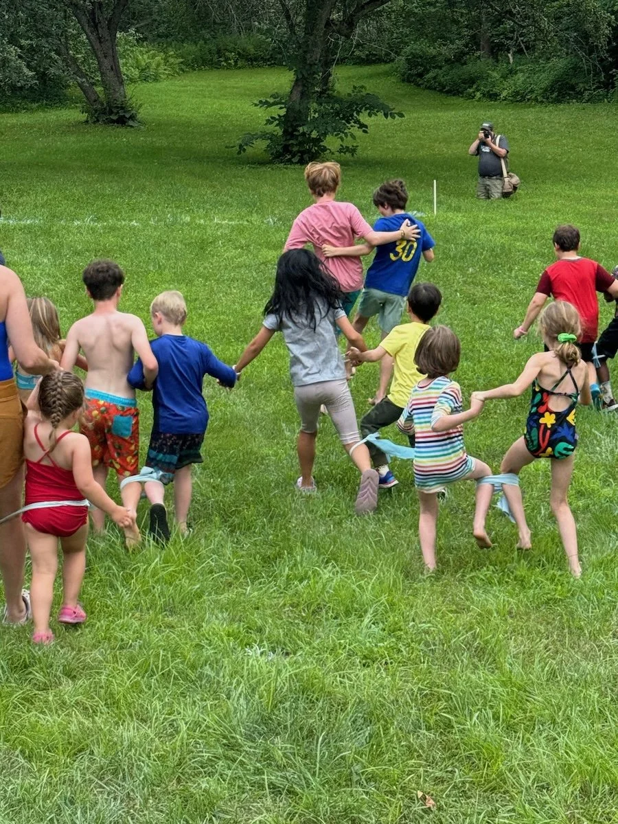 Children and adults are playing a tug-of-war game outside on a grassy field surrounded by trees.