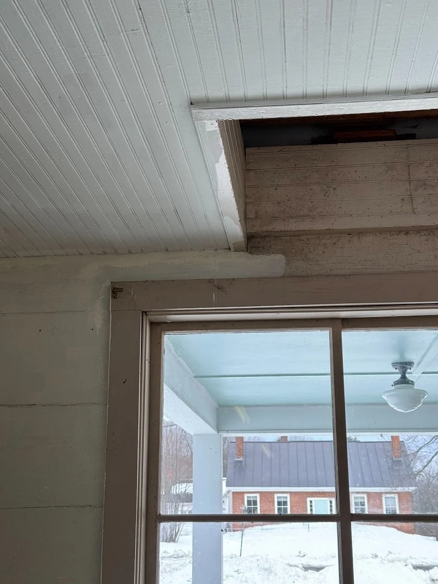 Interior view of a room under renovation, showing a partially exposed ceiling with wood and drywall, a large window with a view of snow-covered ground and a brick house outside.