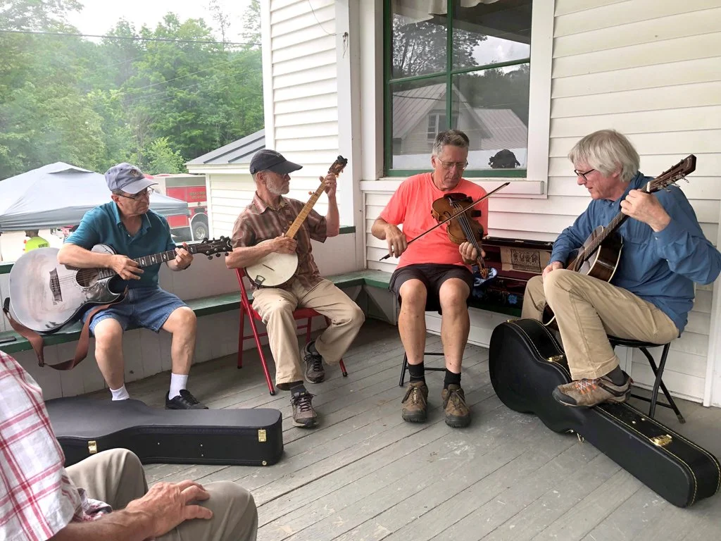 Four men playing musical instruments on a porch, including two with guitars, one with a banjo, and one with a violin.