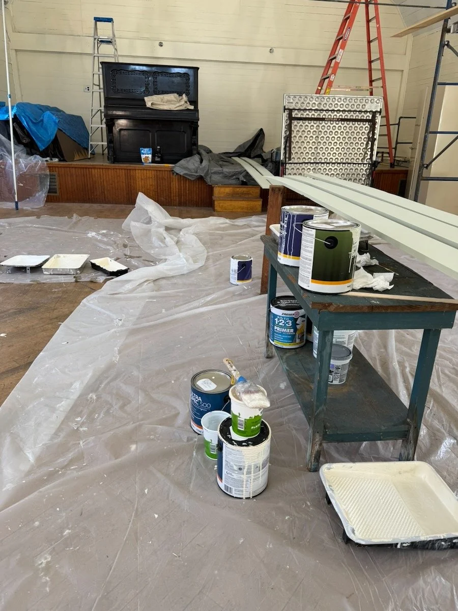 A room under renovation or painting with paint cans, brushes, a plastic sheet on the floor, and scaffolding.