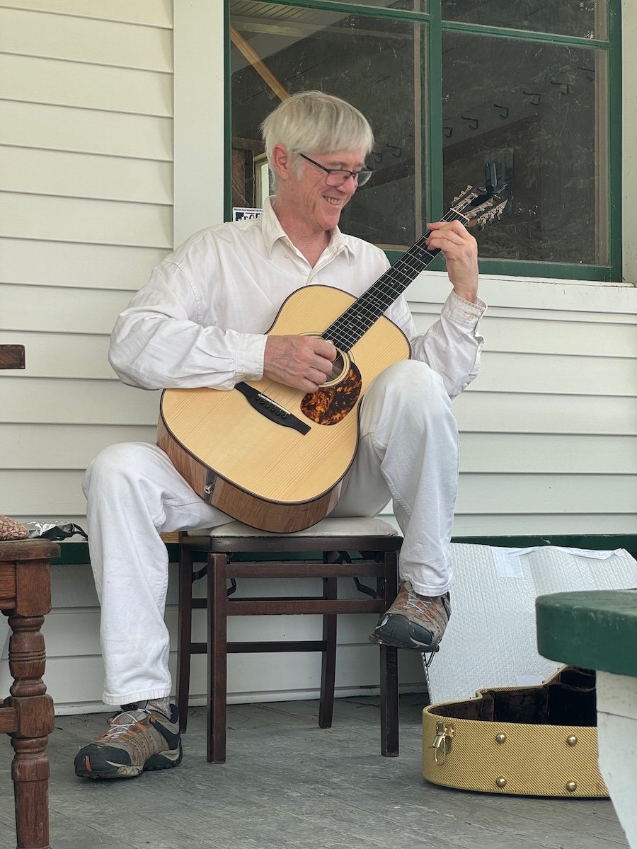 An older man with gray hair, glasses, wearing a white shirt and white pants, is sitting on a chair on a porch playing an acoustic guitar, smiling. A yellow guitar case is on the ground nearby.