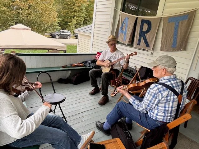 Three people playing musical instruments on a porch with a banner that reads 'ART' in the background.