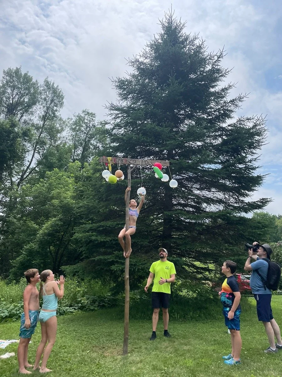Children and adults watching a girl climbing a tall pole decorated with balloons and a sign reading 'Please for' during an outdoor summer event.