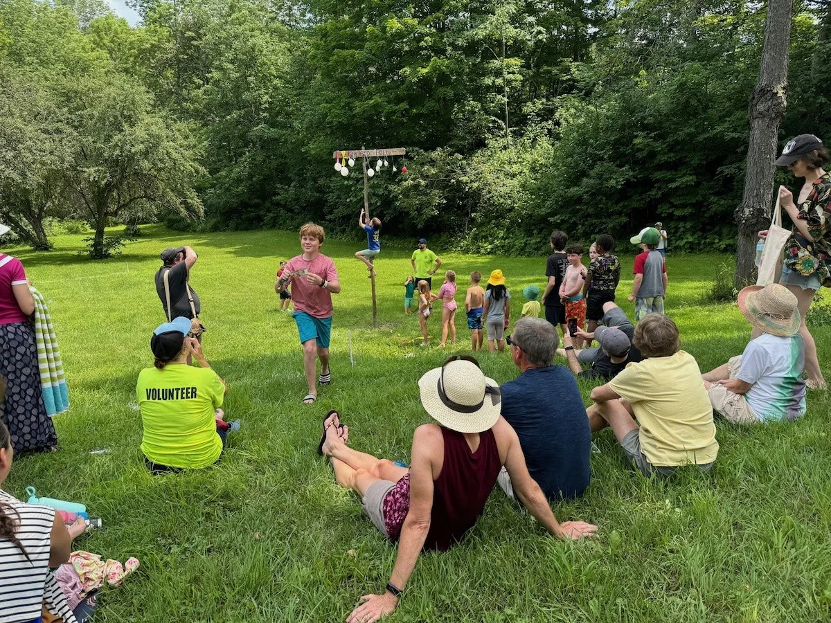 Children and adults gathered outdoors on a grassy area with tall trees in the background, watching a boy running in a race while others participate or observe the event.
