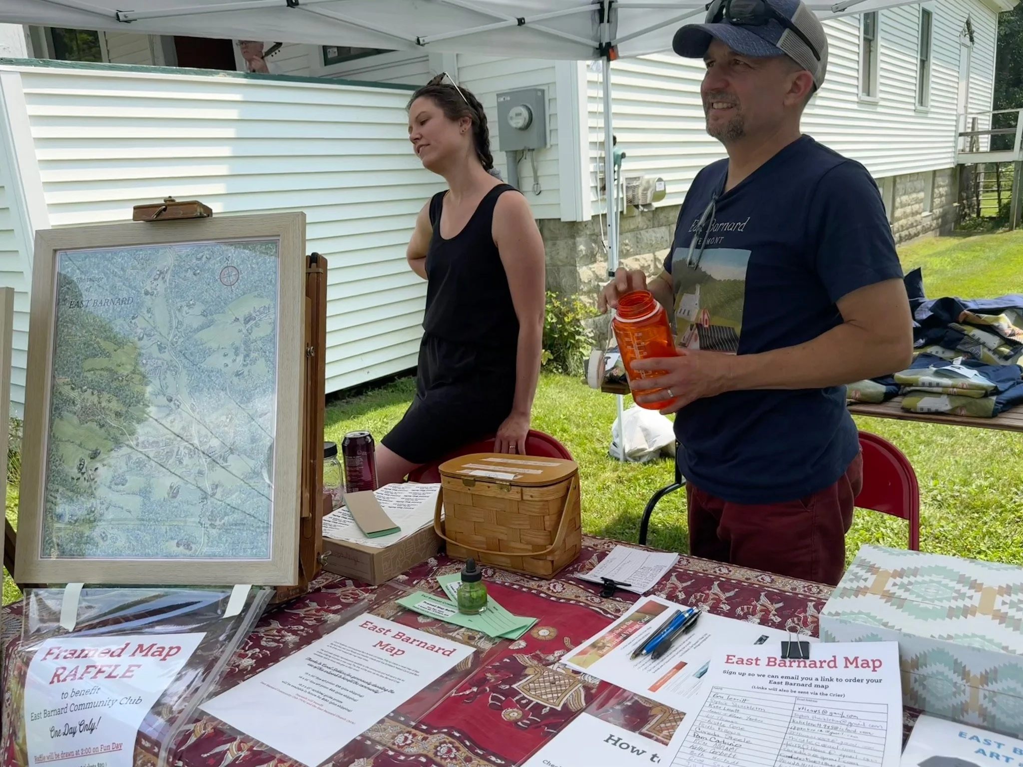 People standing behind a table with maps, flyers, and sign-up sheets at an outdoor event. A man is holding an orange water bottle, and a woman is looking at a display board. A white house with horizontal siding is in the background.