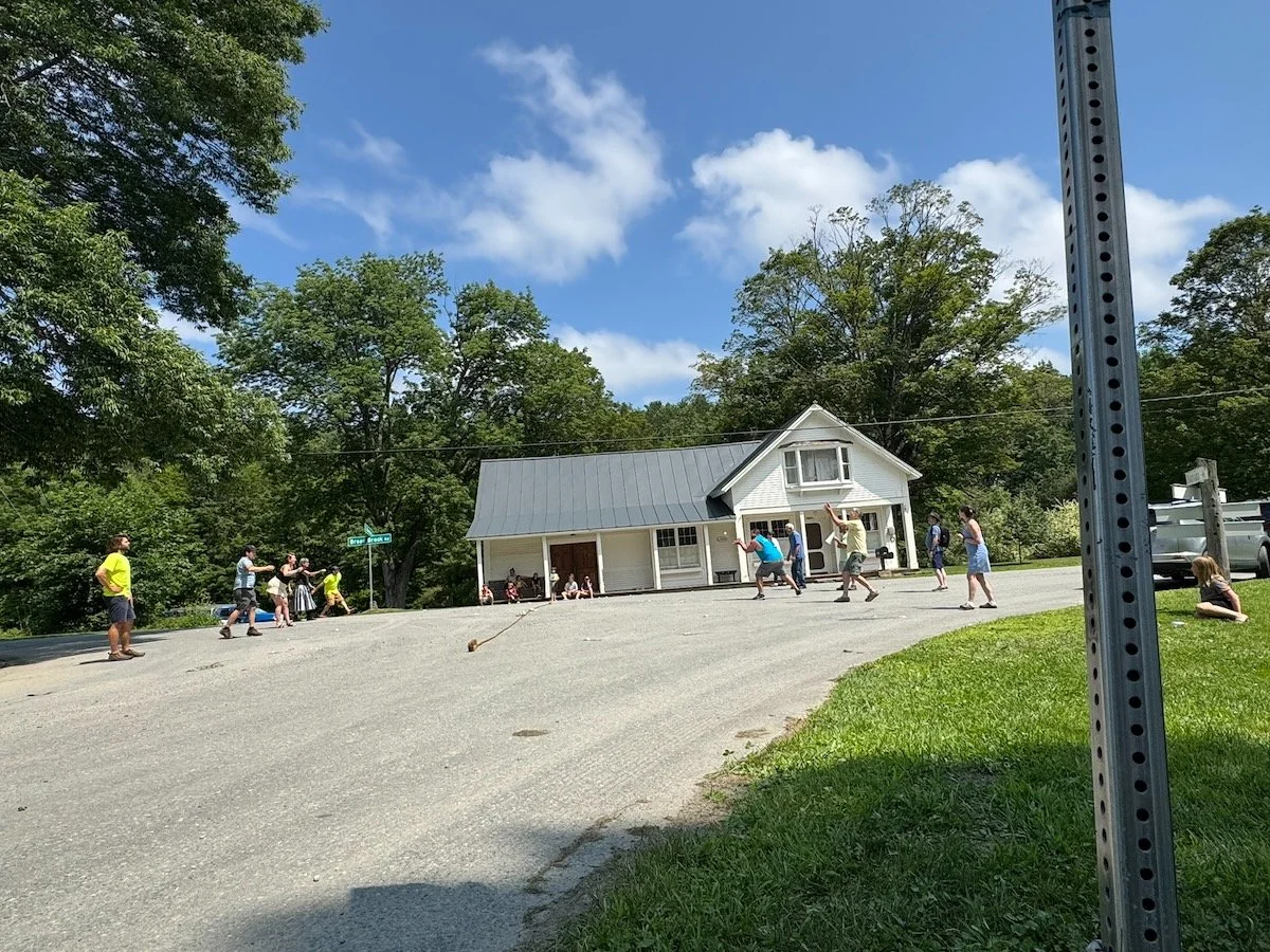 People playing tug of war outside a building with white siding and a gray roof, on a sunny day with a blue sky and some clouds, surrounded by trees and grass.