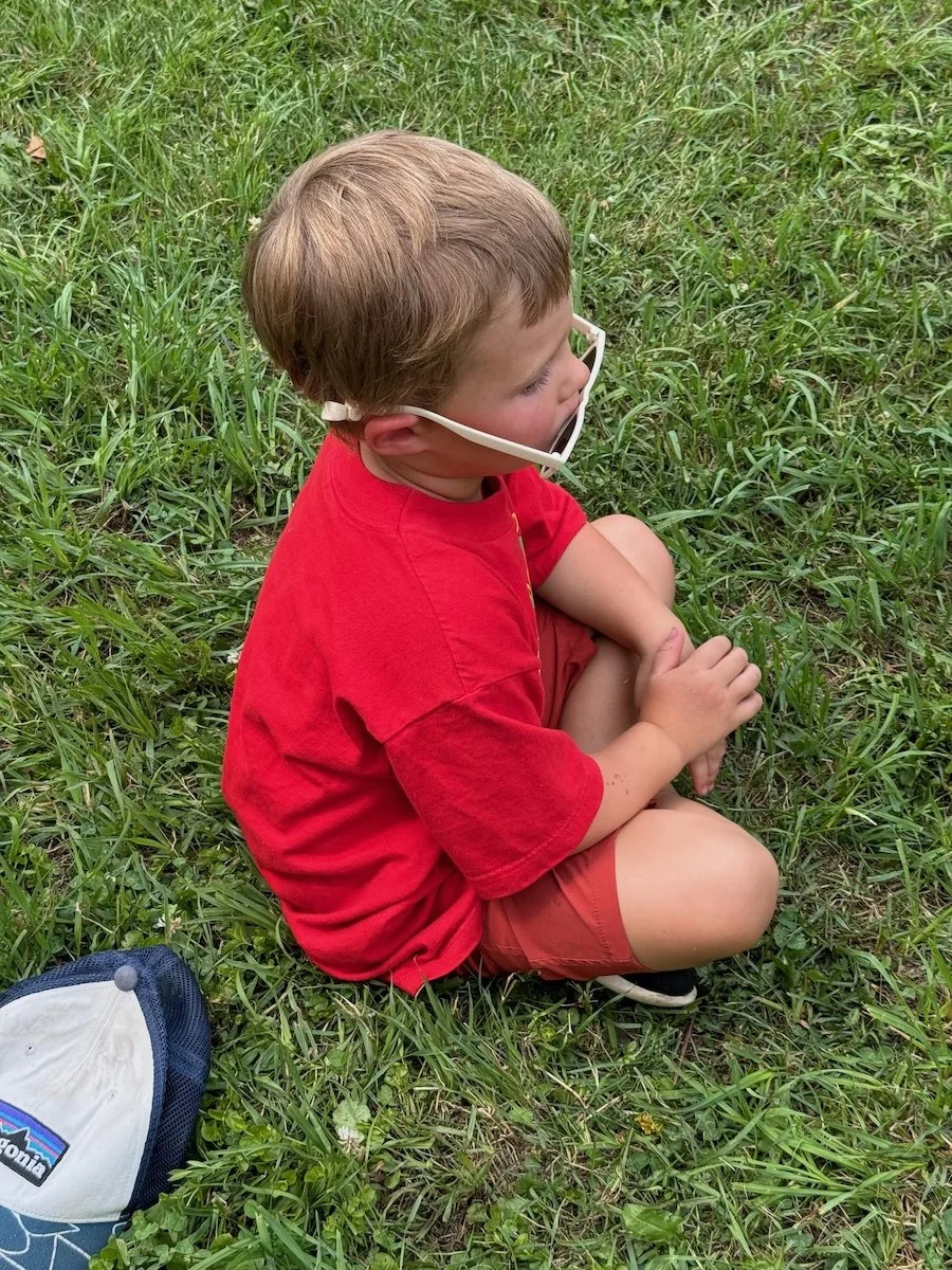 A young boy wearing a red t-shirt and shorts sitting cross-legged on grass, wearing glasses with a strap, outdoors.