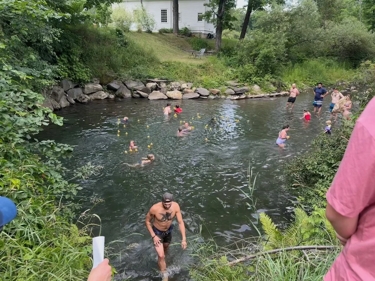 People swimming and wading in a creek surrounded by greenery and rocks with a white house in the background.