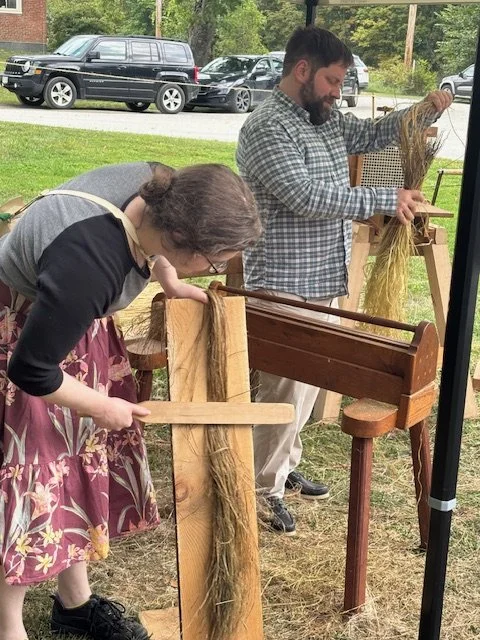 A woman and a man are dyeing wool using traditional hand-dyeing frames outdoors. The woman is holding a wool skein and a wooden paddle, while the man is lifting a bundle of dyed wool. They are standing on grass with parked cars and trees in the backg