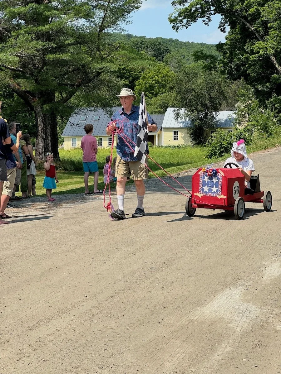 Child in a go-kart decorated with patriotic items, being pulled by an adult with a checkered flag, during a parade with spectators watching on a dirt road surrounded by trees and houses.