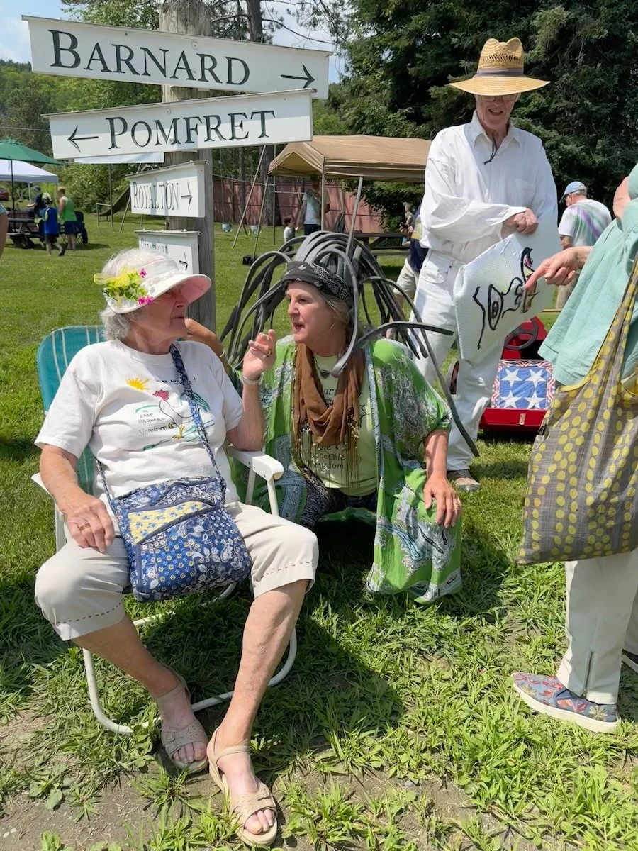 A group of elderly women and a man at an outdoor event, with one woman sitting in a chair with a sun hat and floral decoration, another woman talking to her, both wearing casual clothing and hats. The man, standing with a wide-brimmed hat and white s