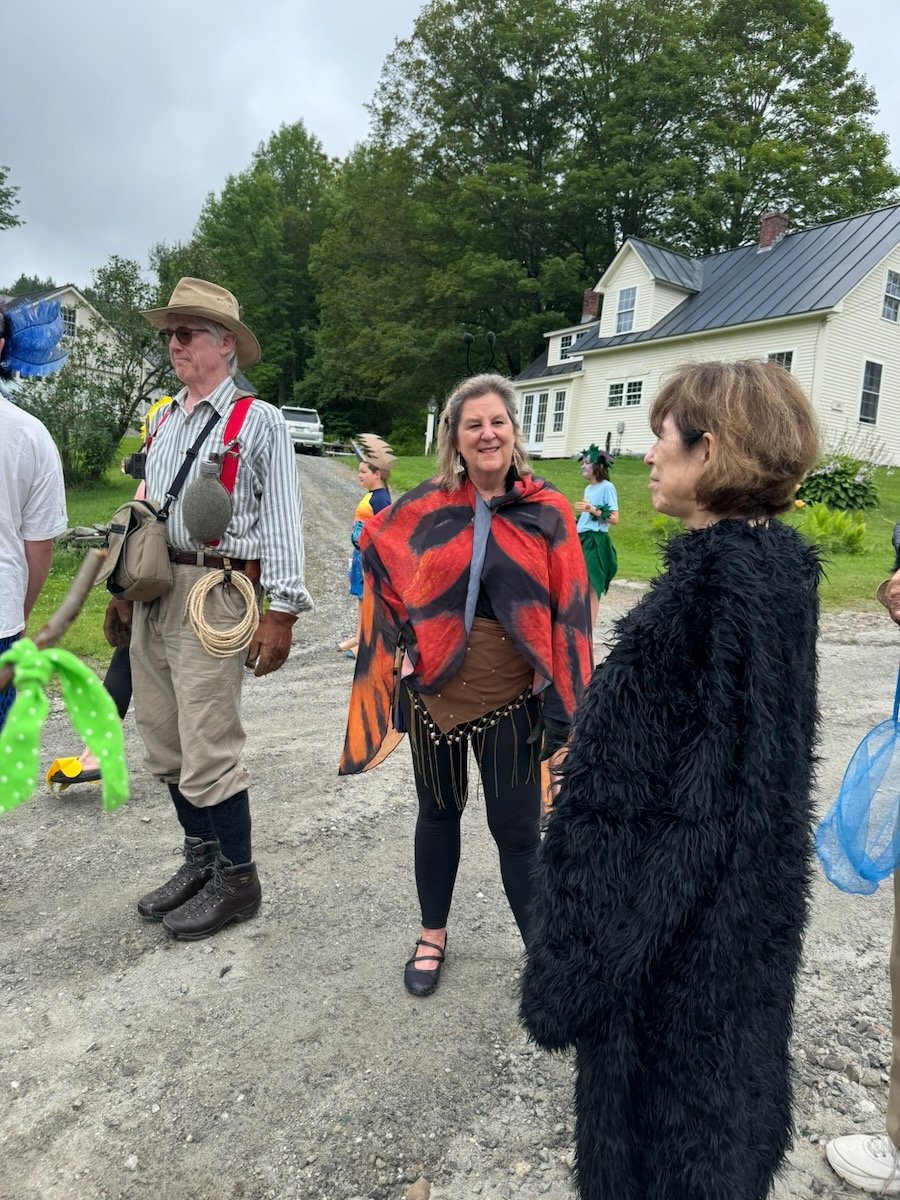 People dressed in costumes and casual clothes standing outdoors, with houses and trees in the background. One woman is wearing a black fuzzy costume, another woman has a red and black butterfly or insect-themed cape, and a man is dressed in outdoor g