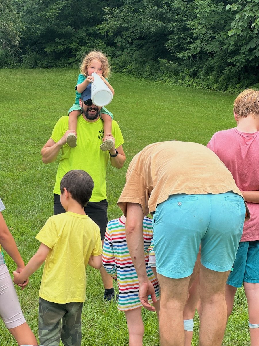 A group of children and adults holding hands outside on a grassy field, with a man carrying a young girl on his shoulders. The girl is drinking from a white cup and has curly blonde hair. The man is wearing a bright yellow shirt and sunglasses, and t