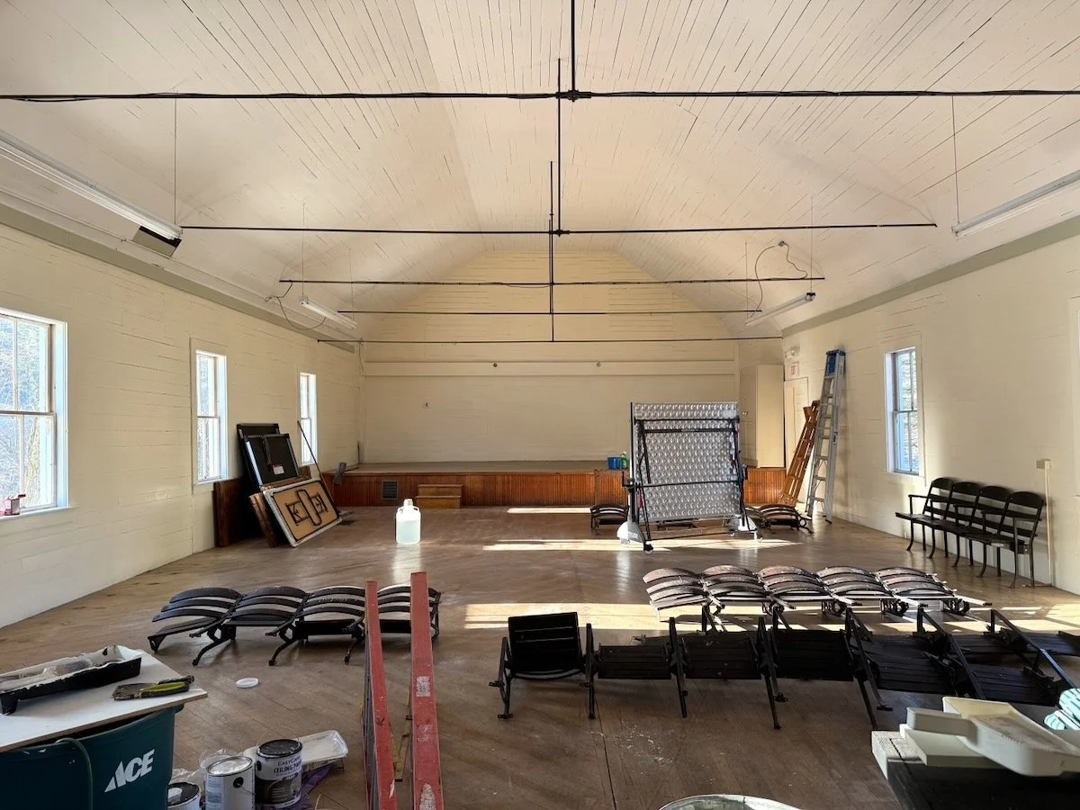 Large empty room with a high arched ceiling, multiple windows, and various chairs stacked and arranged around the floor. Construction tools and materials are scattered around, indicating ongoing setup or renovation.