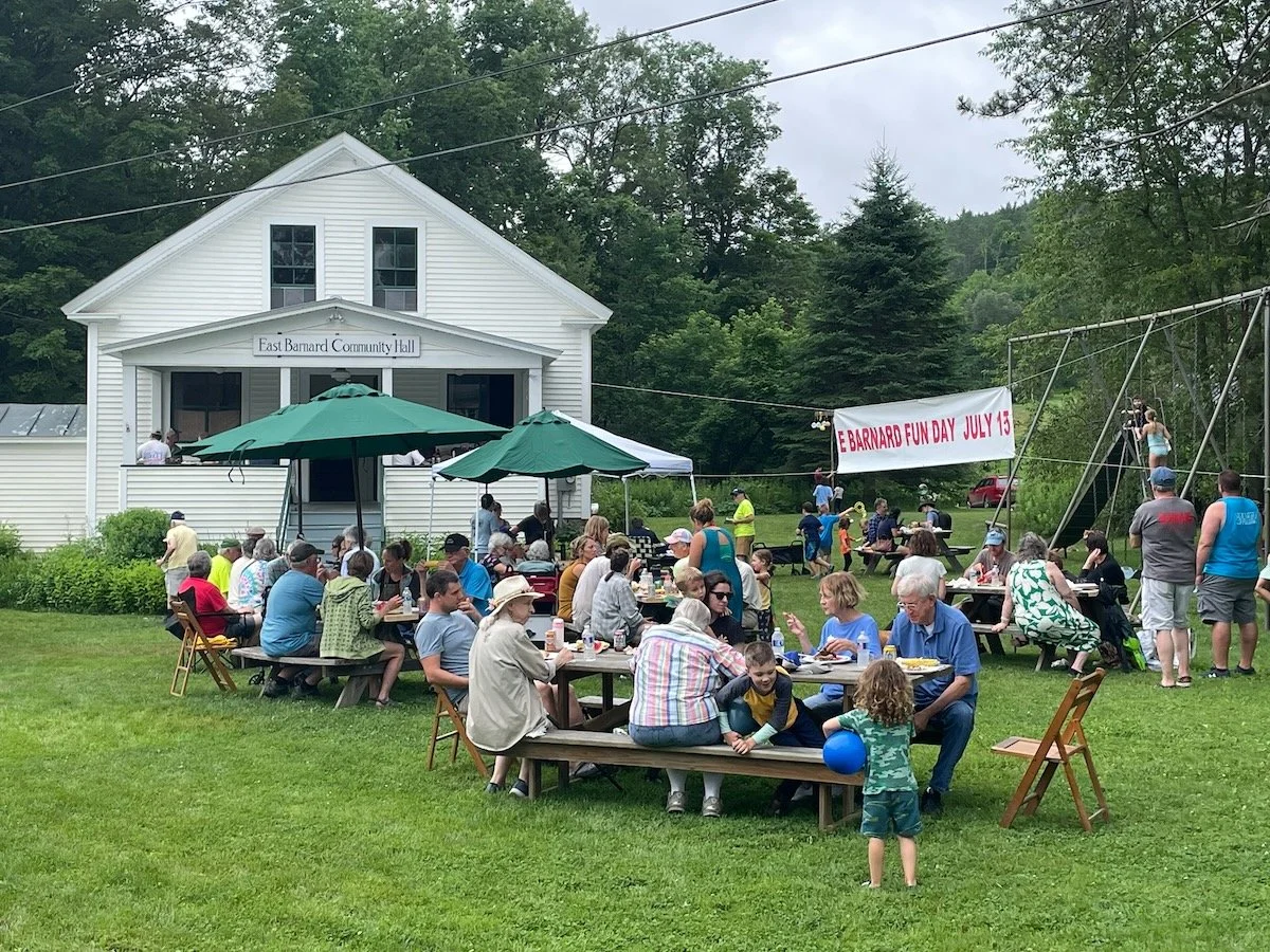 Community gathering on a grassy lawn in front of East Barnard Community Hall, with people sitting at picnic tables enjoying food, children playing, and a banner reading 'East Barnard Fun Day July 15' hanging on a swing set.