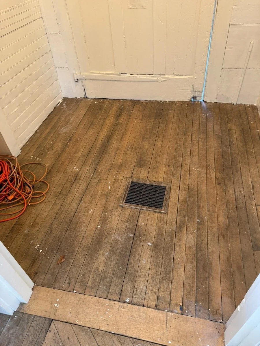 Wooden floor with a floor vent, some scattered dirt and debris, and a pile of orange extension cords in the corner.