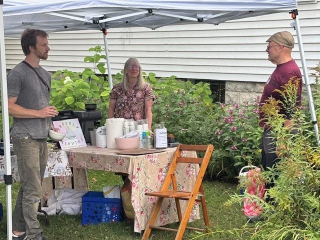 Three people under a white canopy tent, conversing beside a table with supplies, outside in a garden area with bushes and plants.