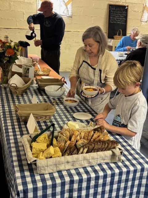 People serving and preparing food at a buffet table covered with a blue and white checkered tablecloth, with bread, cakes, soups, and other dishes.