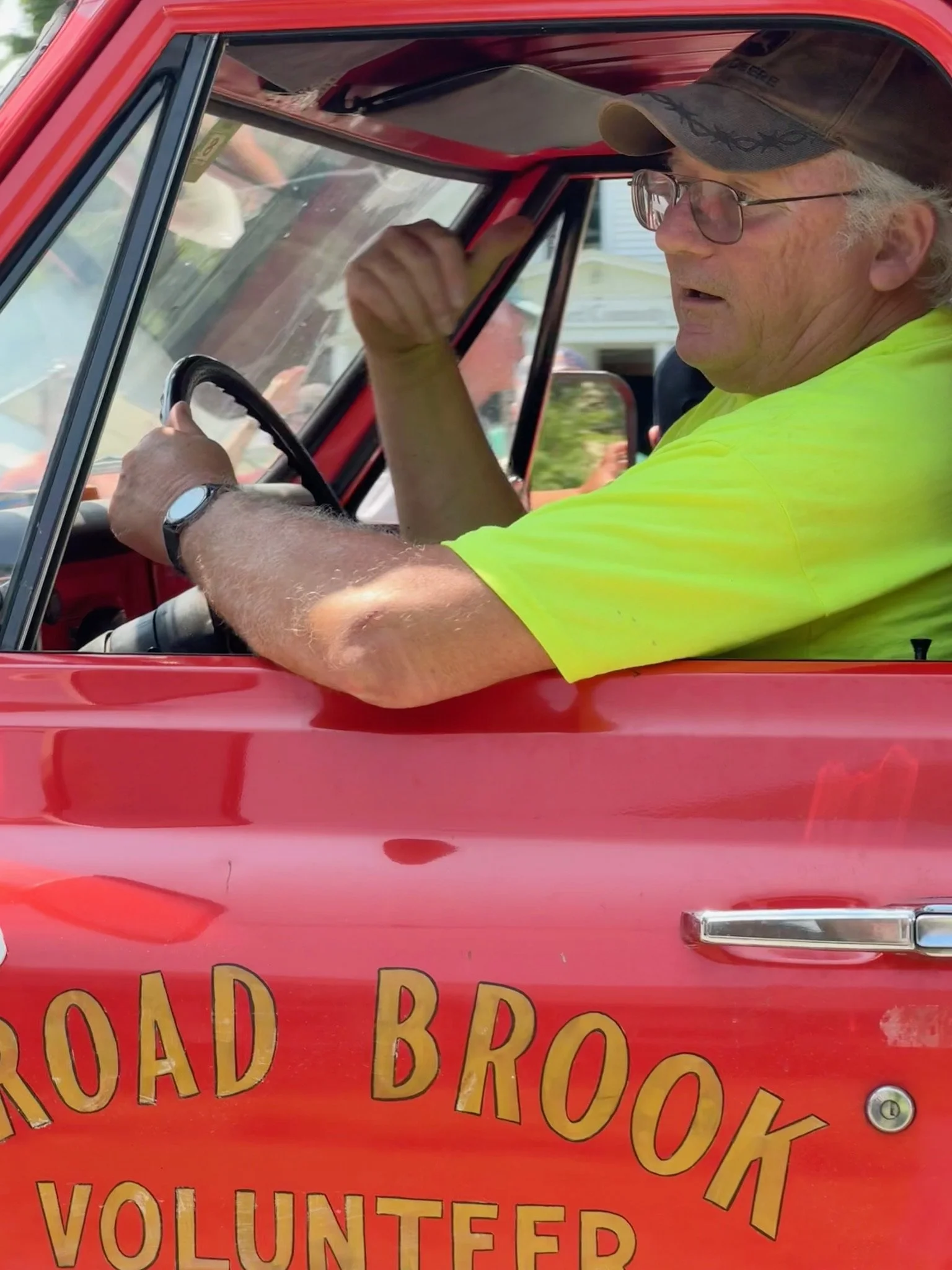 A man wearing a gray baseball cap, glasses, and a bright yellow shirt is sitting inside a red food truck with the words "ROAD BROOK VOLUNTEER" written on the side.