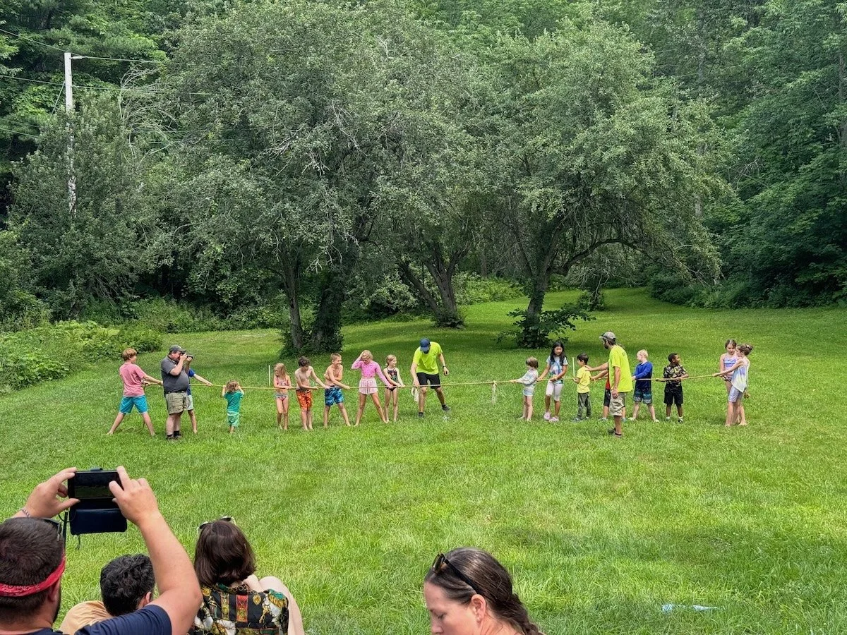 Children and adults participating in a tug-of-war game on a grassy field surrounded by trees, with spectators watching and taking photos.
