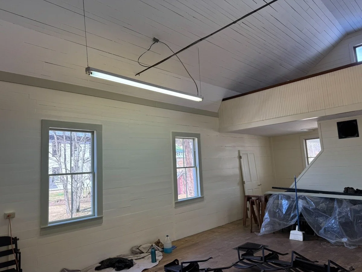 Interior of a room with white walls and ceiling, two windows showing trees outside, and some furniture covered with plastic. There is a fluorescent light fixture hanging from the ceiling.